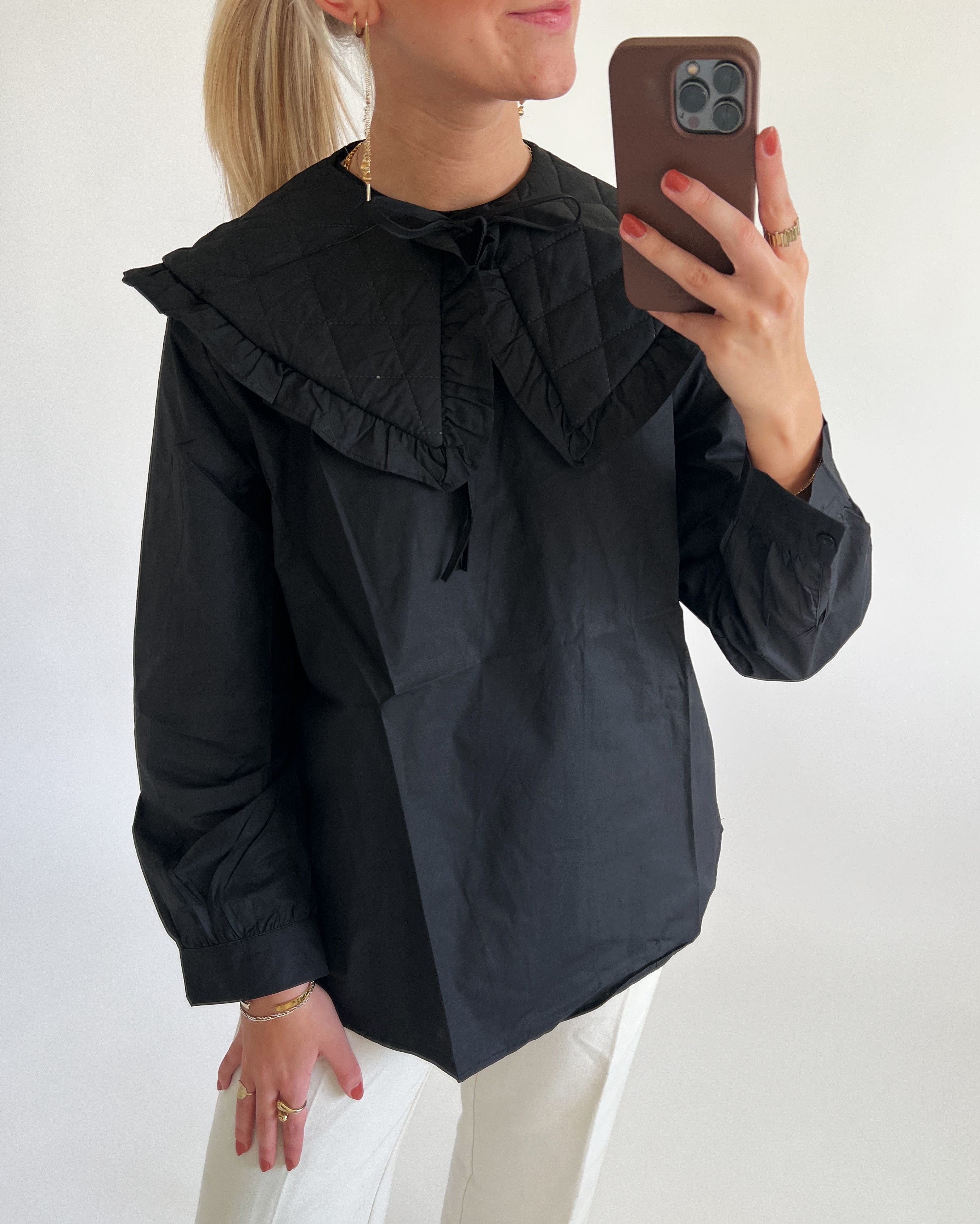 En person, iført sort bluse med stor krave, tager en selfie i spejlet med en mobiltelefon. Personen bærer også øreringe og flere ringe, og har lyst hår.