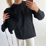 En person iført sort skjorte tager en selfie med en smartphone, mens de holder en sort quiltet jakke. Baggrunden er en ensfarvet, lys væg.