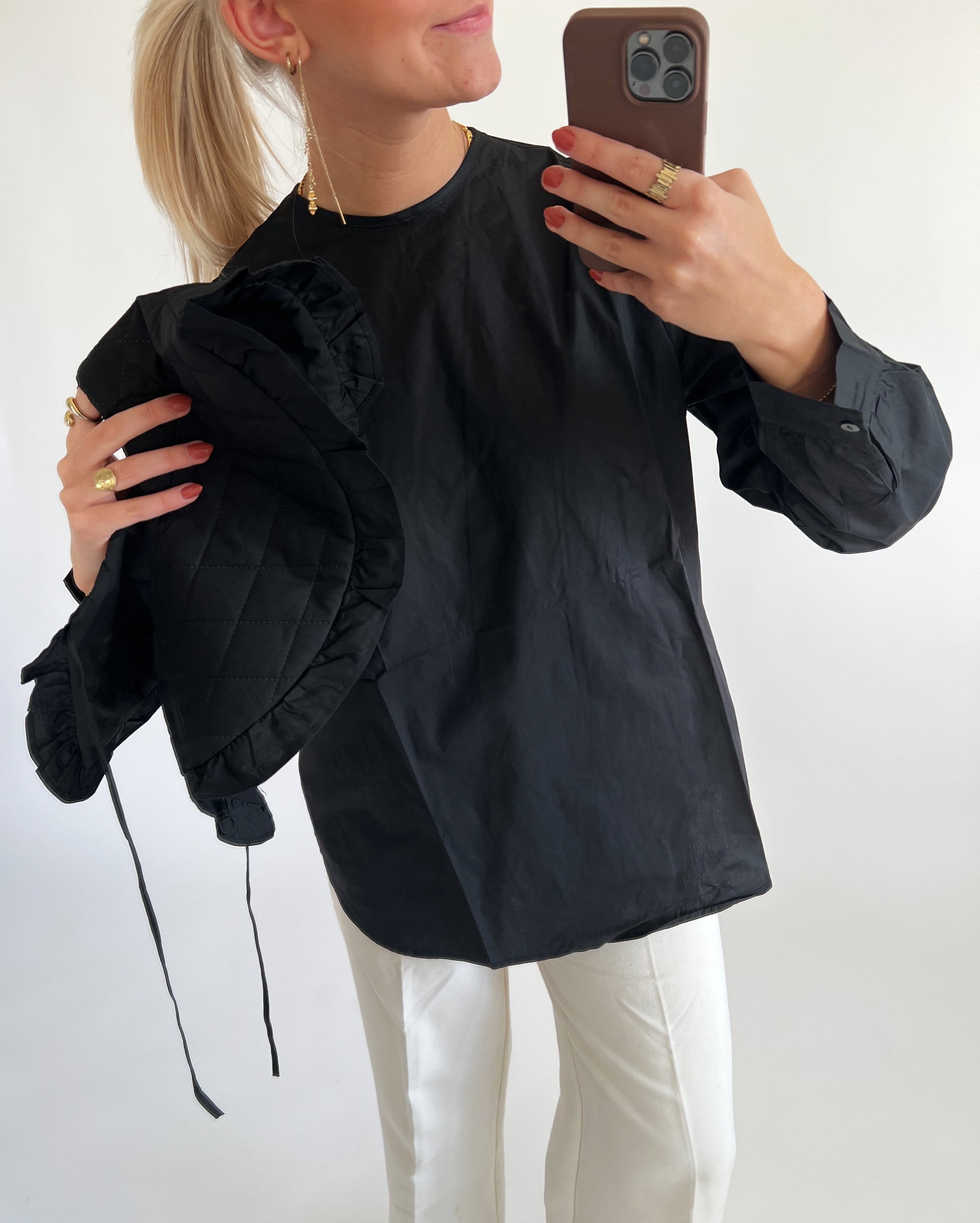 En person iført sort skjorte tager en selfie med en smartphone, mens de holder en sort quiltet jakke. Baggrunden er en ensfarvet, lys væg.