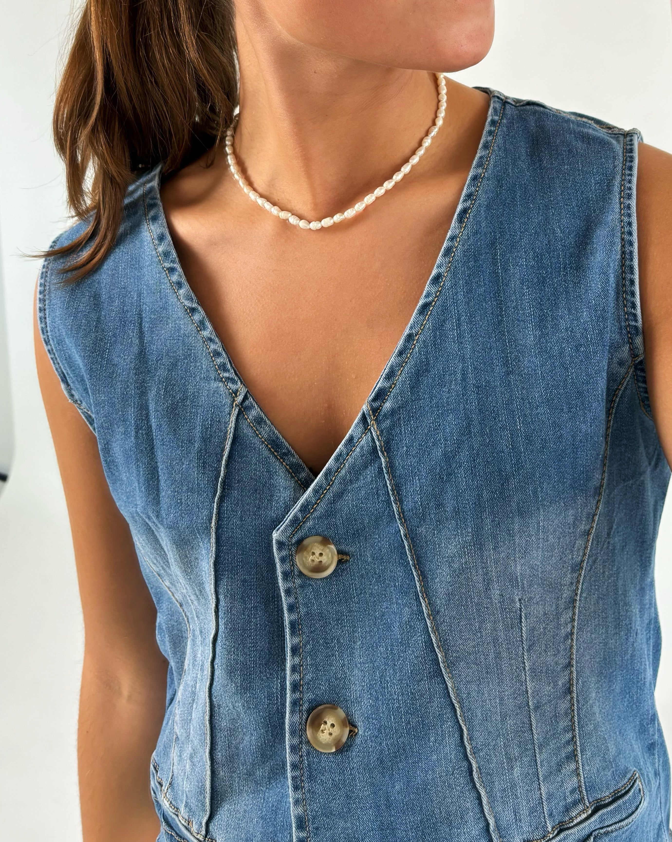 En person iført en ærmeløs denimvest med tre knapper og en perlekæde rundt om halsen står mod en lys baggrund.