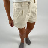 En persons underkrop med cremefarvede shorts og grå sko står på hvid baggrund. Personen bærer et hvidt ribbet tanktop og smykker på venstre hånd.