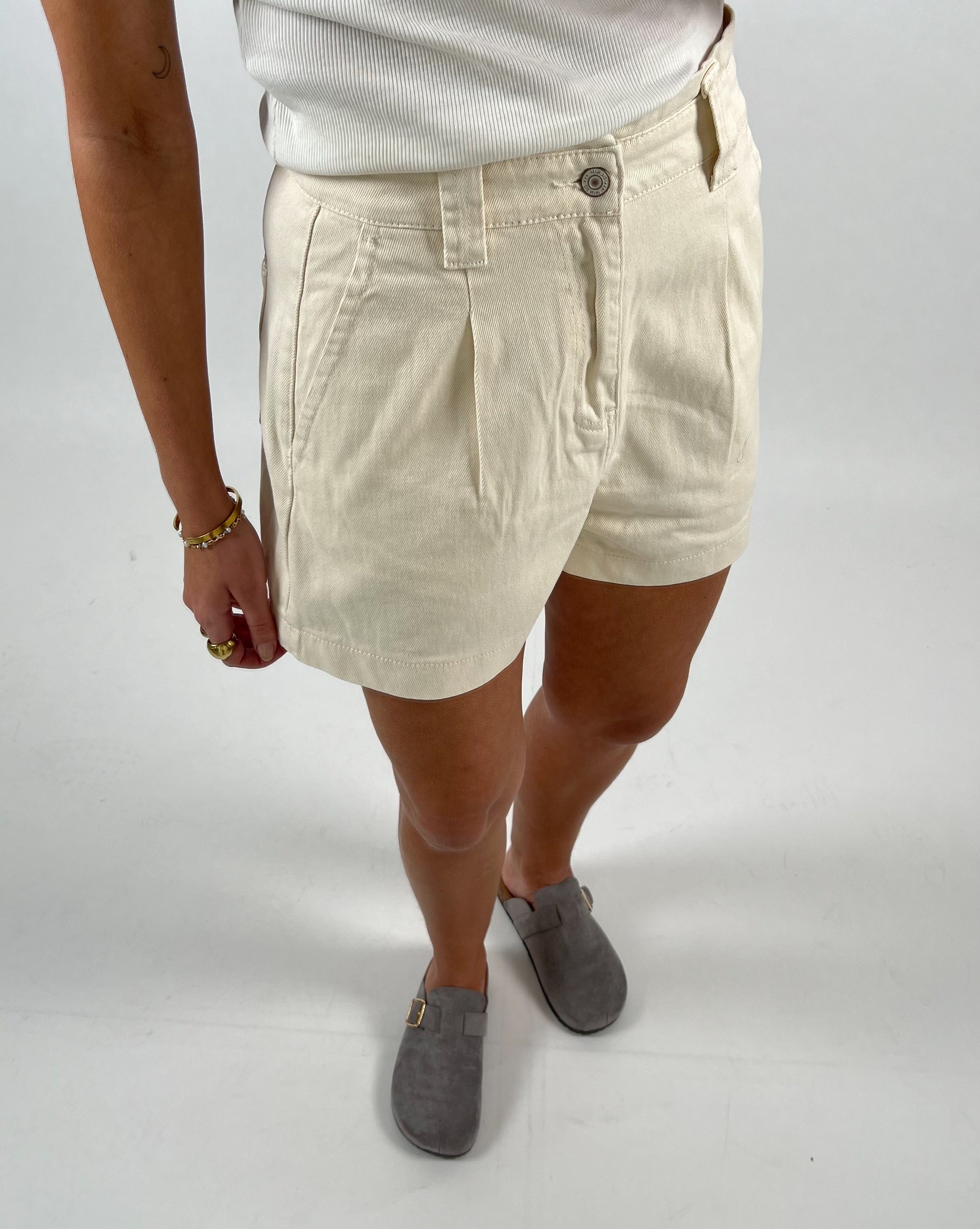 En persons underkrop med cremefarvede shorts og grå sko står på hvid baggrund. Personen bærer et hvidt ribbet tanktop og smykker på venstre hånd.