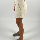 En person iført beige shorts står med siden til, iført grå hjemmesko. Personen har en hvid top på, og der ses en arm med armbånd. Baggrunden er hvid.
