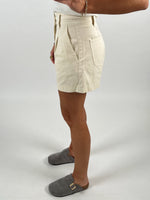 En person iført beige shorts står med siden til, iført grå hjemmesko. Personen har en hvid top på, og der ses en arm med armbånd. Baggrunden er hvid.