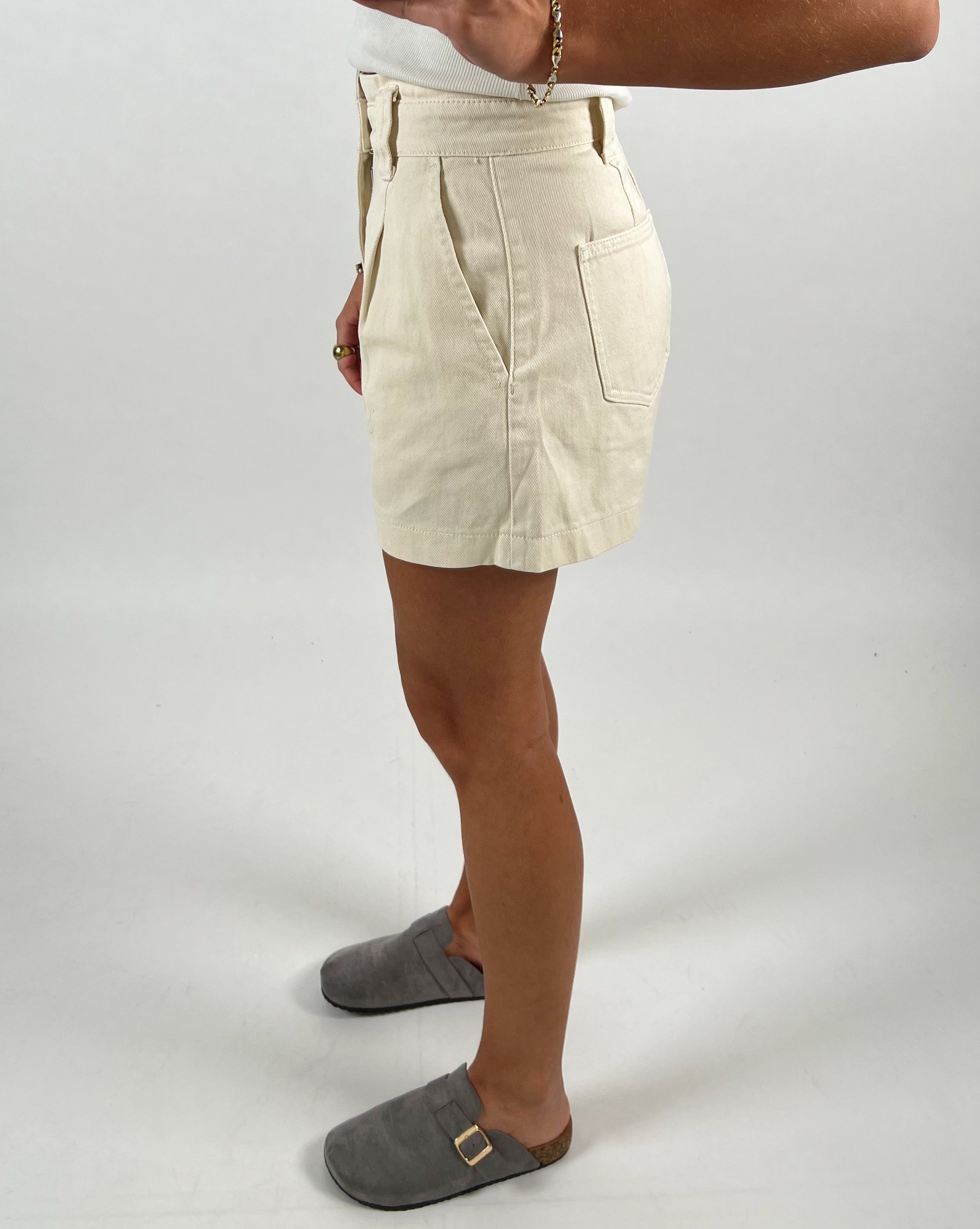 En person iført beige shorts står med siden til, iført grå hjemmesko. Personen har en hvid top på, og der ses en arm med armbånd. Baggrunden er hvid.