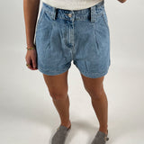 En person iført denimshorts og en hvid top står i grå loafers. Personen bærer armbånd og en ring på højre hånd. Baggrunden er enkel og hvid.