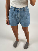 En person iført denimshorts og en hvid top står i grå loafers. Personen bærer armbånd og en ring på højre hånd. Baggrunden er enkel og hvid.