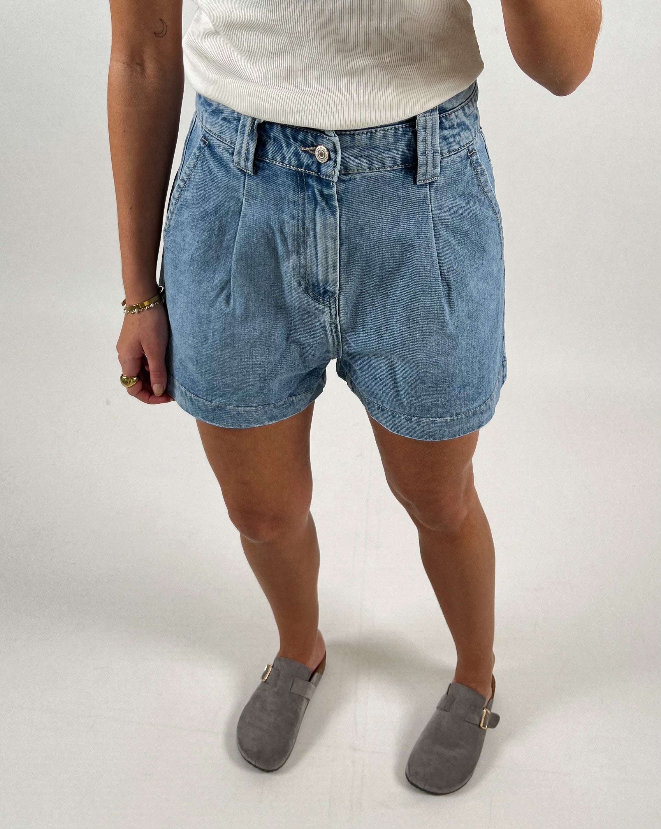 En person iført denimshorts og en hvid top står i grå loafers. Personen bærer armbånd og en ring på højre hånd. Baggrunden er enkel og hvid.