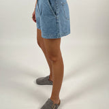 En person iført denimshorts står i en profilvisning mod en neutral, hvid baggrund. Personen bærer grå tøfler, og deres overkrop er delvist uden for rammen.