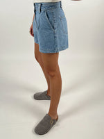 En person iført denimshorts står i en profilvisning mod en neutral, hvid baggrund. Personen bærer grå tøfler, og deres overkrop er delvist uden for rammen.