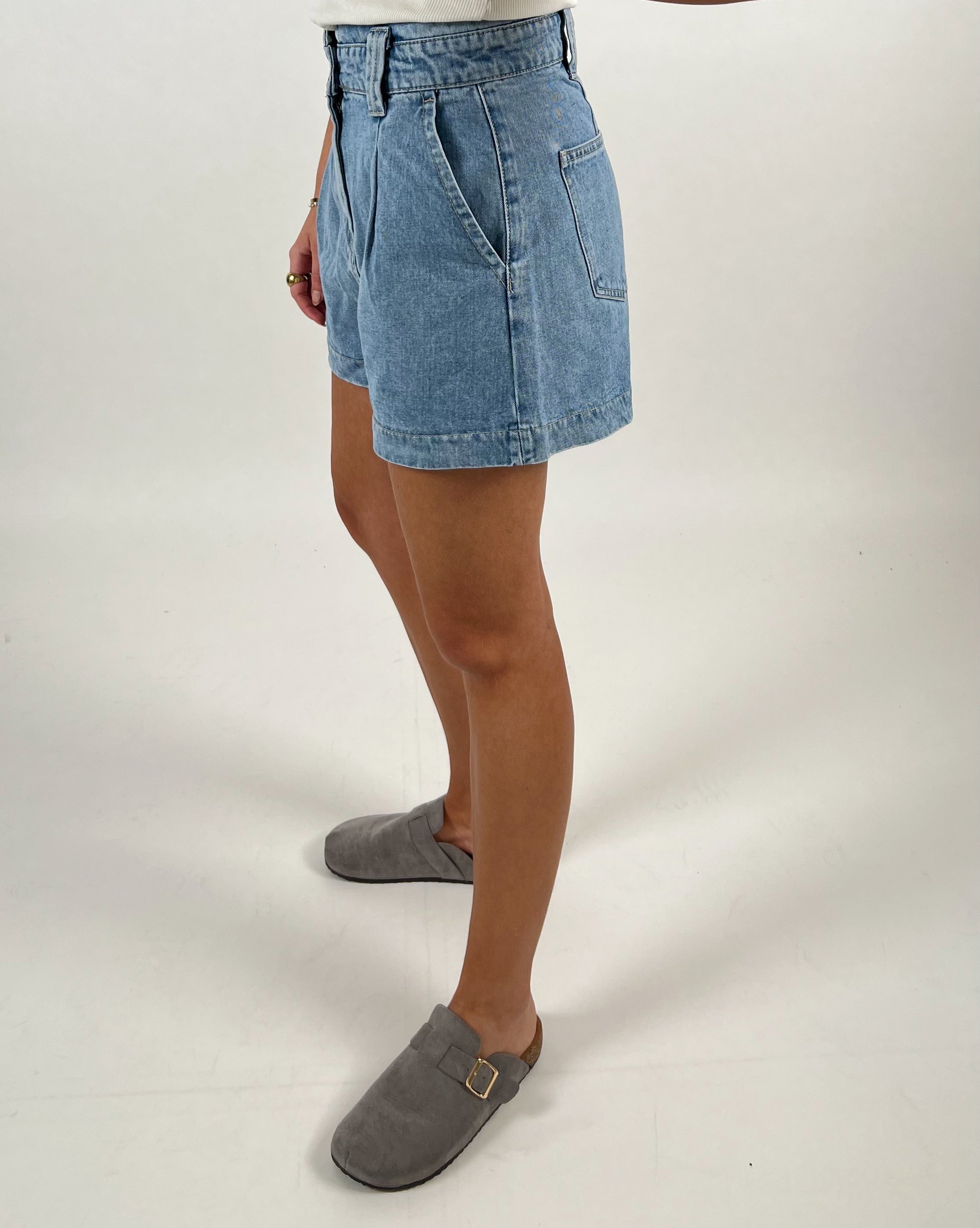 En person iført denimshorts står i en profilvisning mod en neutral, hvid baggrund. Personen bærer grå tøfler, og deres overkrop er delvist uden for rammen.
