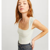 En person med rødt hår bærer en lyseblå tanktop og mørke jeans, står i en let vinklet positur mod en neutral, lys baggrund.