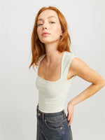 En person med rødt hår bærer en lyseblå tanktop og mørke jeans, står i en let vinklet positur mod en neutral, lys baggrund.