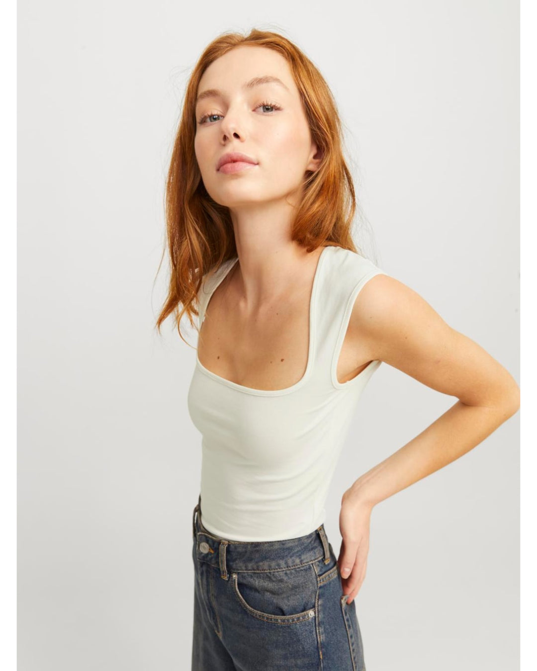 En person med rødt hår bærer en lyseblå tanktop og mørke jeans, står i en let vinklet positur mod en neutral, lys baggrund.