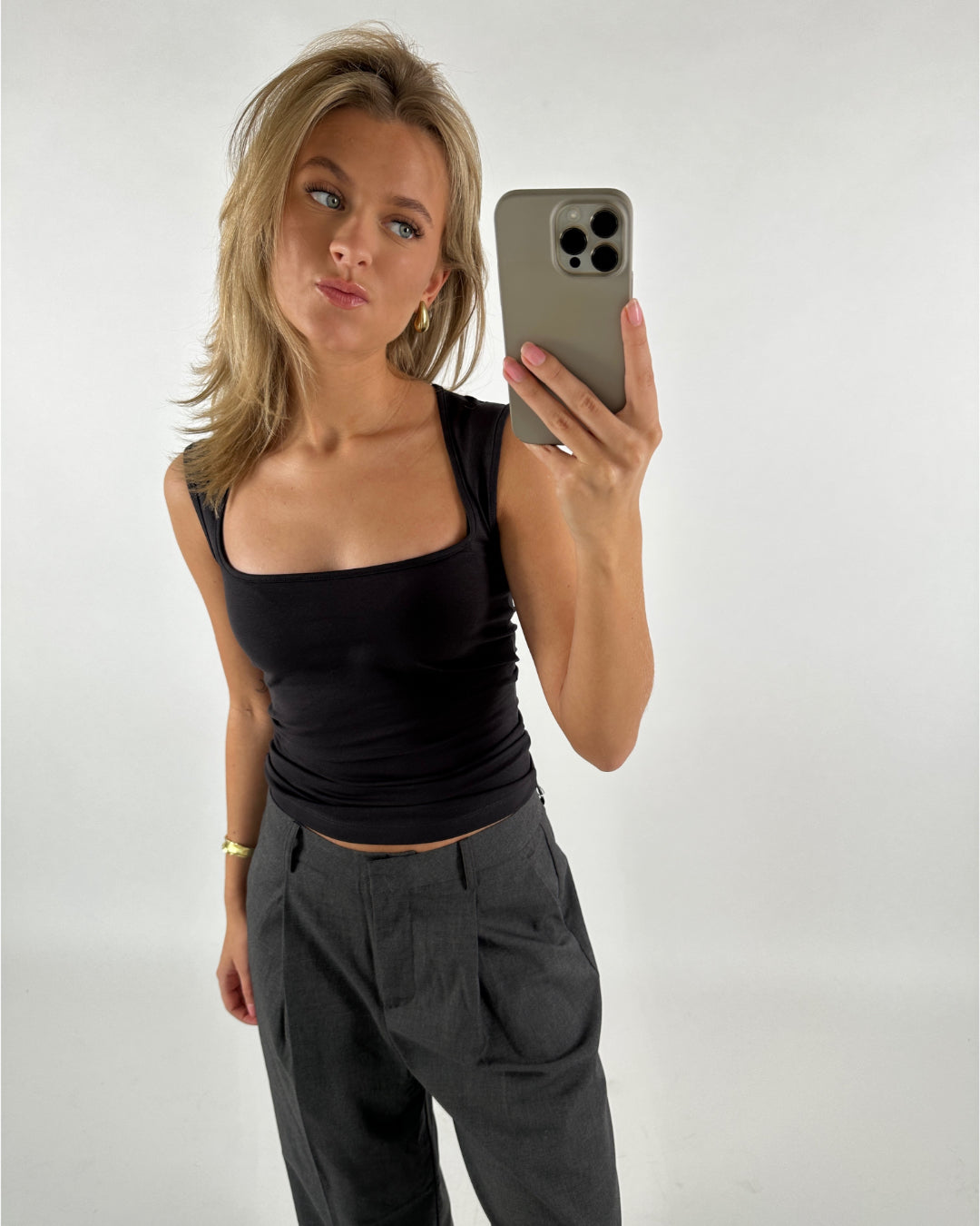 Person står og tager en selfie med en smartphone. De har en sort top og grå bukser på. Baggrunden er simpel og lys, hvilket fremhæver personen.