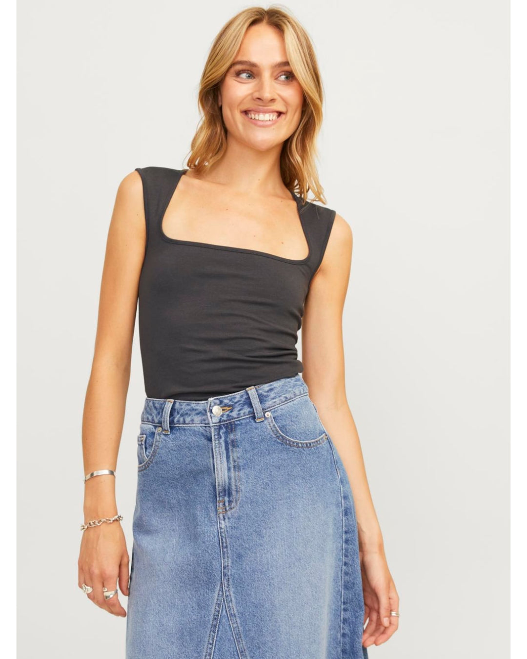 Kvinde smiler iført sort top og denimnederdel, står afslappet i et lyst indendørs miljø. Hun bærer armbånd og ringe, med lang, bølget hår.