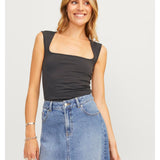 Kvinde smiler iført sort top og denimnederdel, står afslappet i et lyst indendørs miljø. Hun bærer armbånd og ringe, med lang, bølget hår.