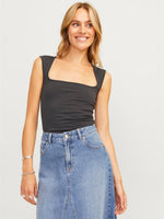 Kvinde smiler iført sort top og denimnederdel, står afslappet i et lyst indendørs miljø. Hun bærer armbånd og ringe, med lang, bølget hår.