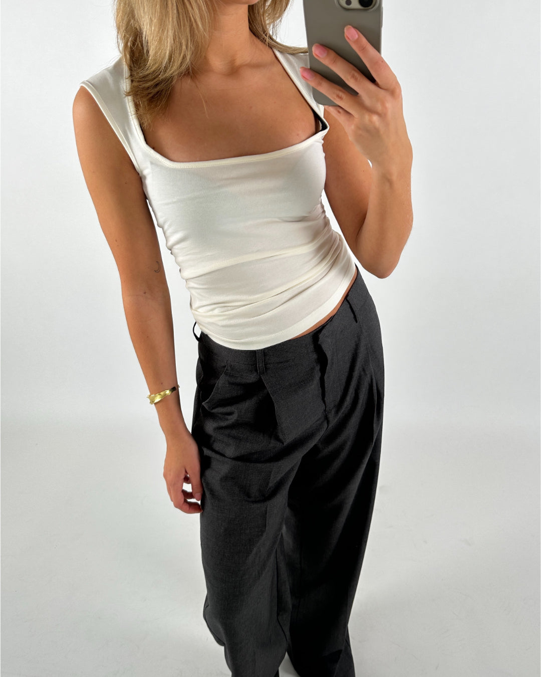 Kvinde iført hvid top og sorte bukser tager et spejlbillede. Hun holder en smartphone og står i et hvidt, neutralt rum. En gylden armbånd pryder hendes venstre arm.