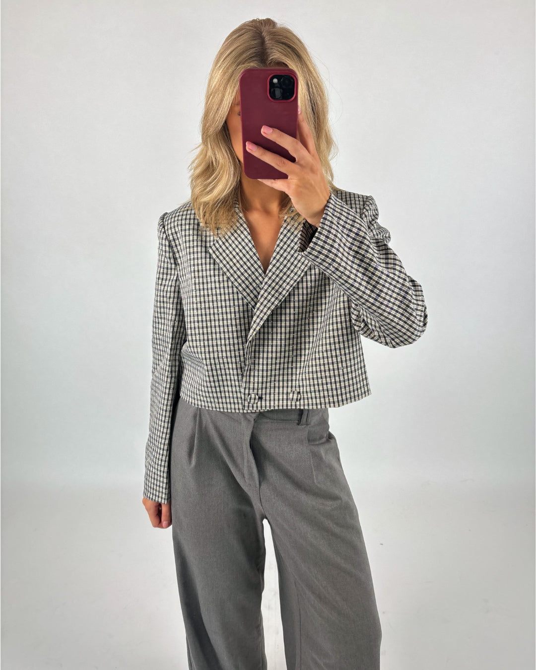 Kvinde holder en telefon med rød etui foran ansigtet, iført en ternet blazer og grå bukser, i et minimalistisk rum med hvid baggrund.