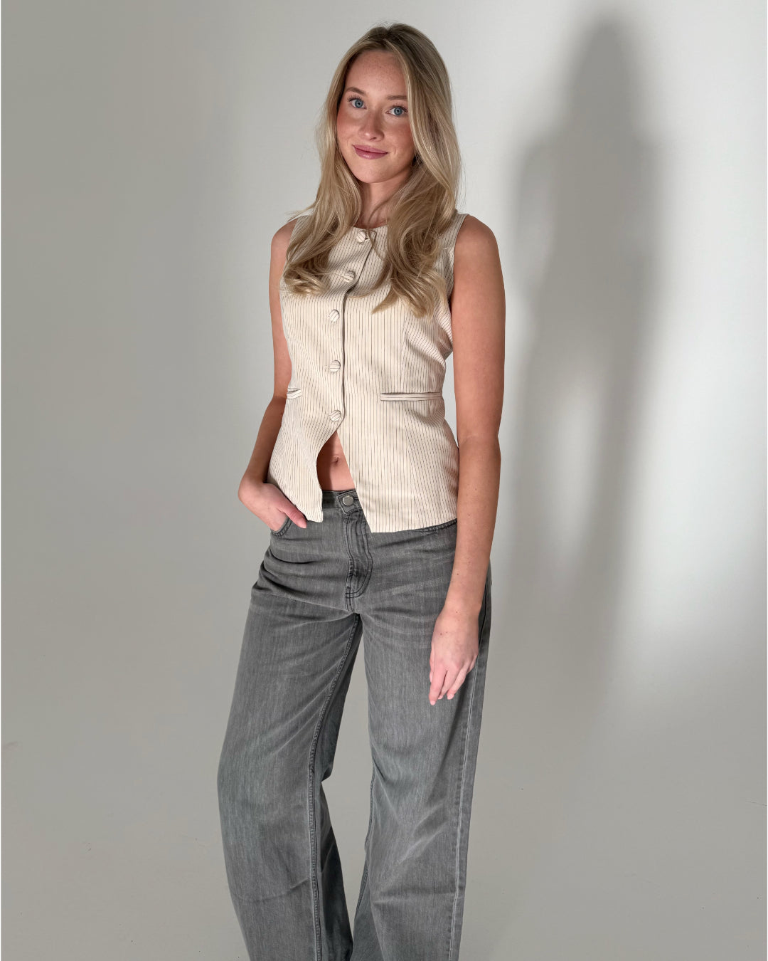 En kvinde står afslappet med den ene hånd i lommen i en neutral indendørs studieindstilling. Hun bærer en beige top med knapper og grå, løse jeans.