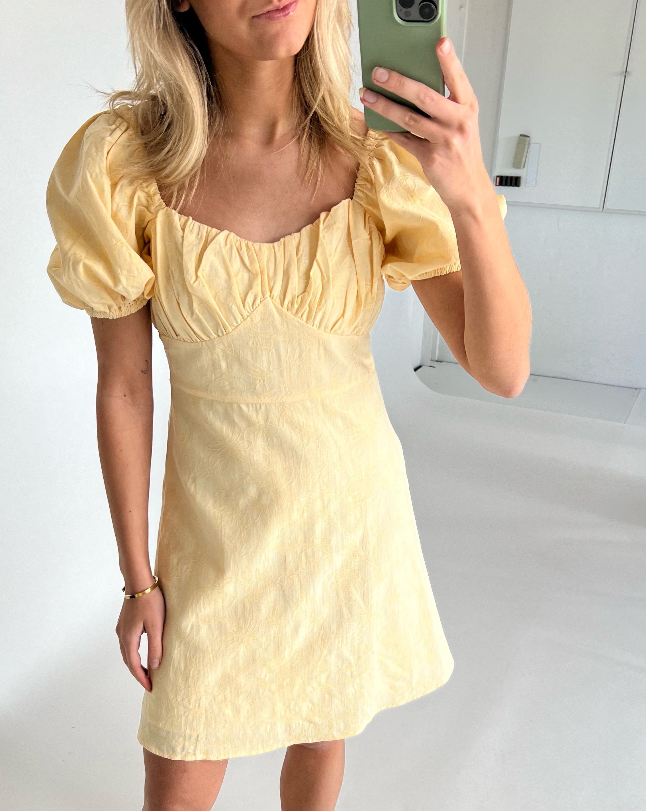 En person iført en gul kjole tager en selfie i et spejl. De står i et lyst rum med hvide vægge og en neutral baggrund.