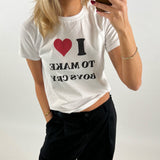 Kvinde iført hvid T-shirt med teksten "I ❤️ TO MAKE BOYS CRY", tager et spejlbillede. Hun bærer sorte bukser og har rød neglelak. Baggrunden er enkel og lys.