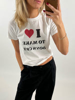 Kvinde iført hvid T-shirt med teksten "I ❤️ TO MAKE BOYS CRY", tager et spejlbillede. Hun bærer sorte bukser og har rød neglelak. Baggrunden er enkel og lys.