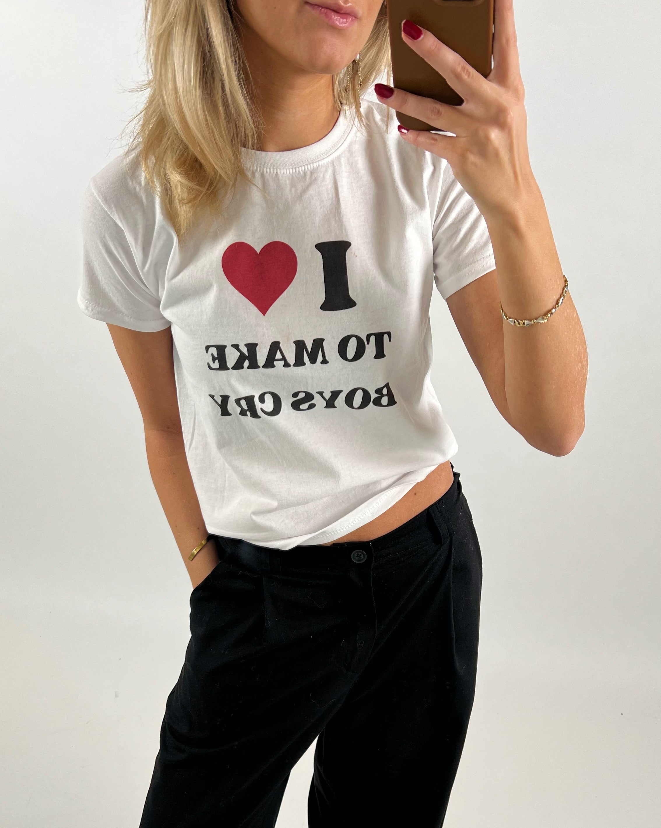 Kvinde iført hvid T-shirt med teksten "I ❤️ TO MAKE BOYS CRY", tager et spejlbillede. Hun bærer sorte bukser og har rød neglelak. Baggrunden er enkel og lys.