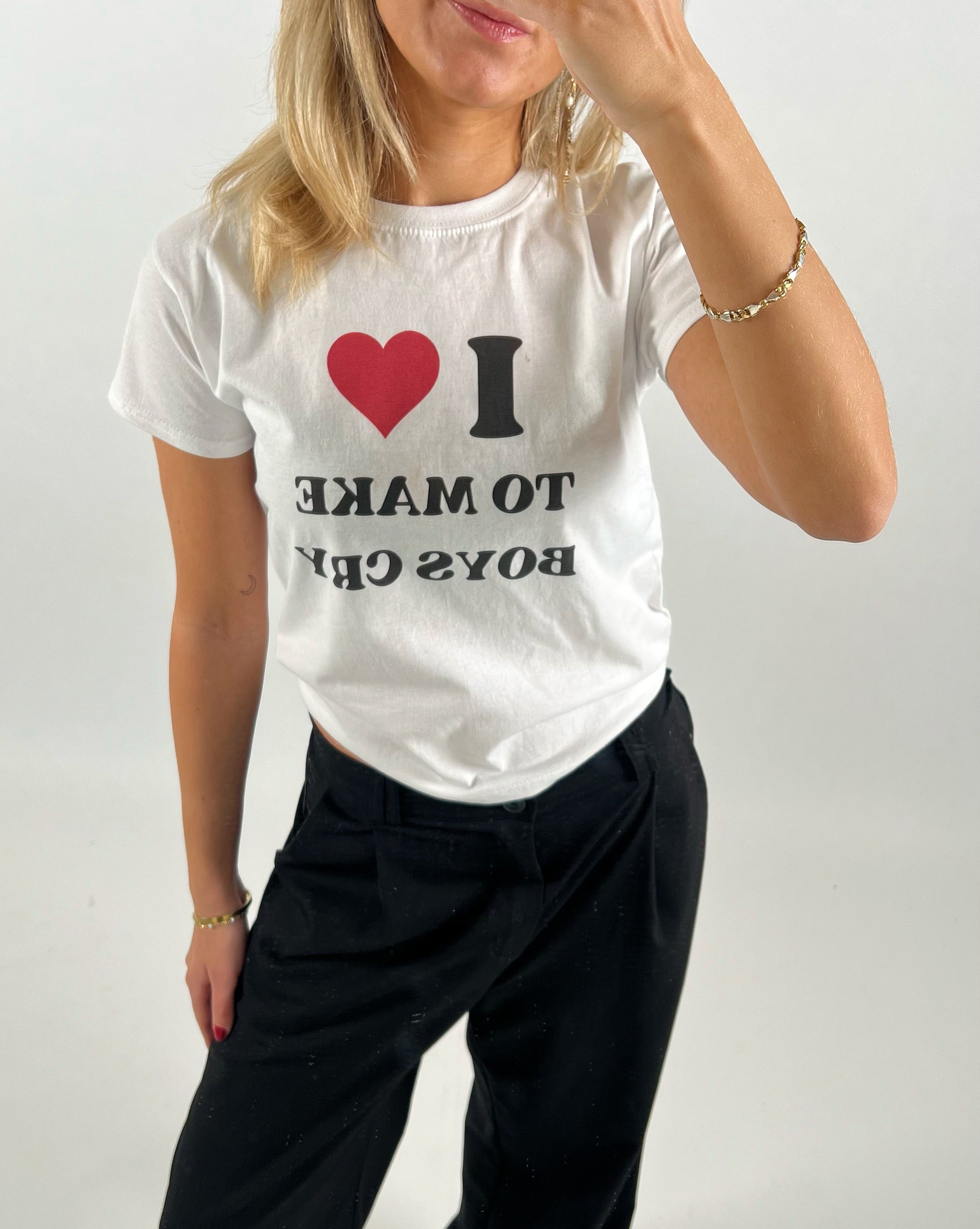 Kvinde bærer en hvid T-shirt med teksten "I ❤️ TO MAKE BOYS CRY" i sort og rød skrift. Hun står i et neutralt rum og dækker delvist ansigtet med hånden.