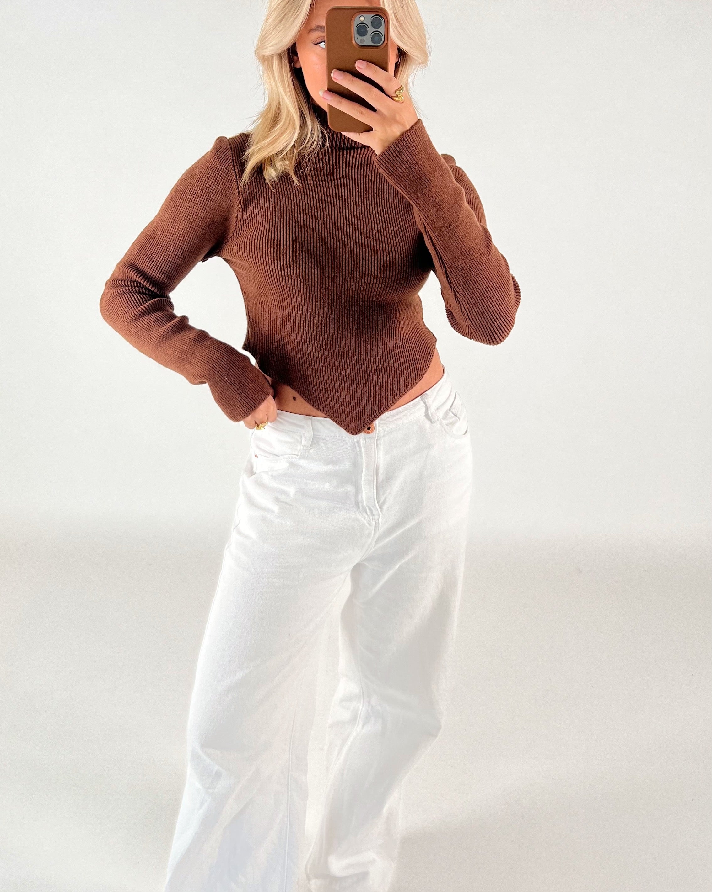En person iført en brun sweater og hvide bukser tager et spejlbillede med en smartphone i et neutralt, hvidt rum.