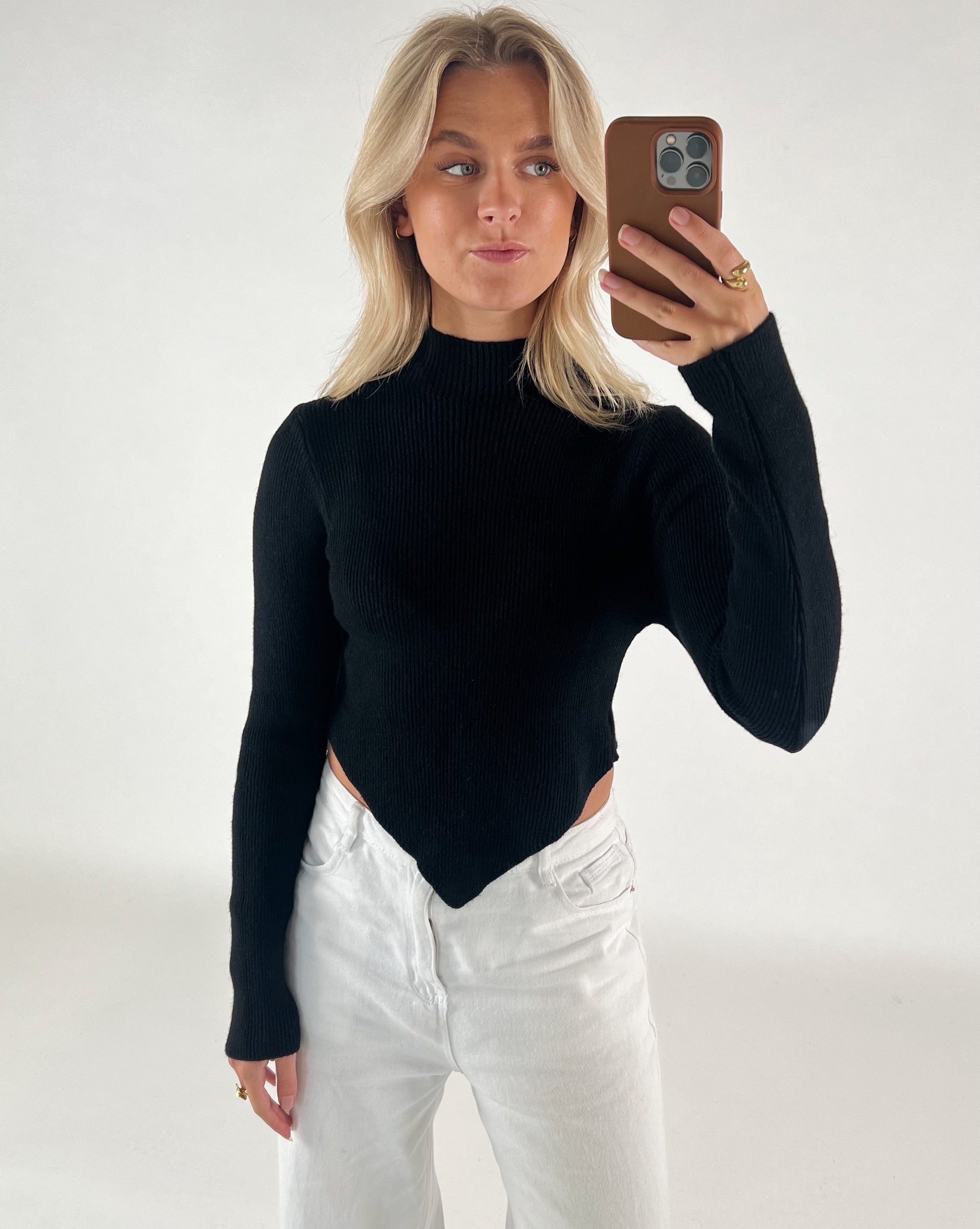 En person tager en selfie i et spejl, iført en sort trøje og hvide bukser. Personen holder en brun telefon, og baggrunden er simpel og lys.
