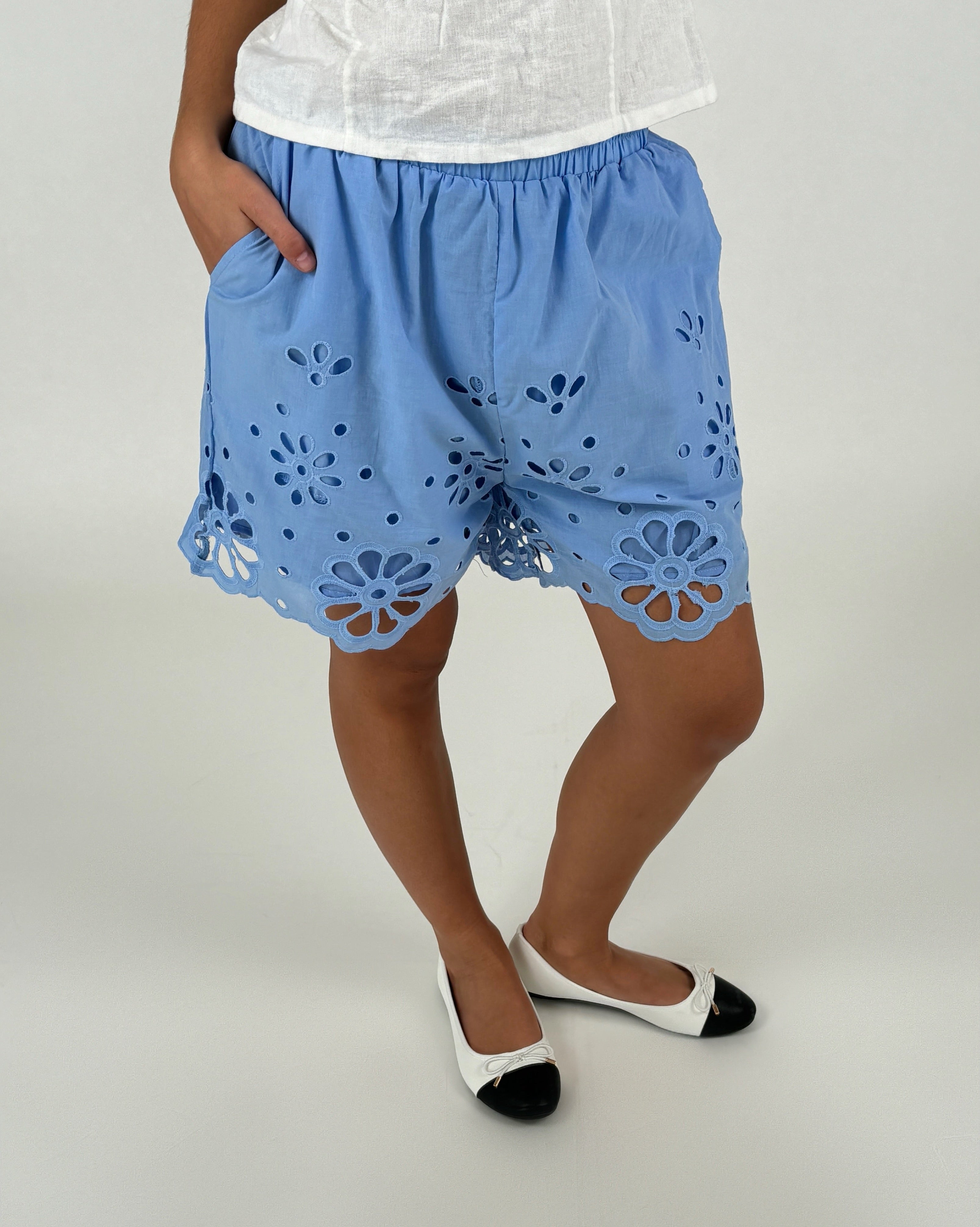 Blå shorts med blomsterudskæringer bæres af en person, der står oprejst. Personen har hænderne i lommerne. En hvid overdel og sko komplementerer det lyse baggrund.