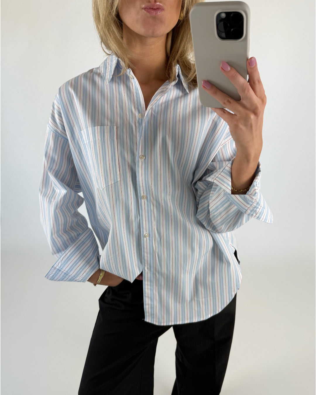 En person iført en stribet skjorte tager et selfiespejl i en minimalistisk indretning, mens de poser og holder en smartphone med en lys baggrund.