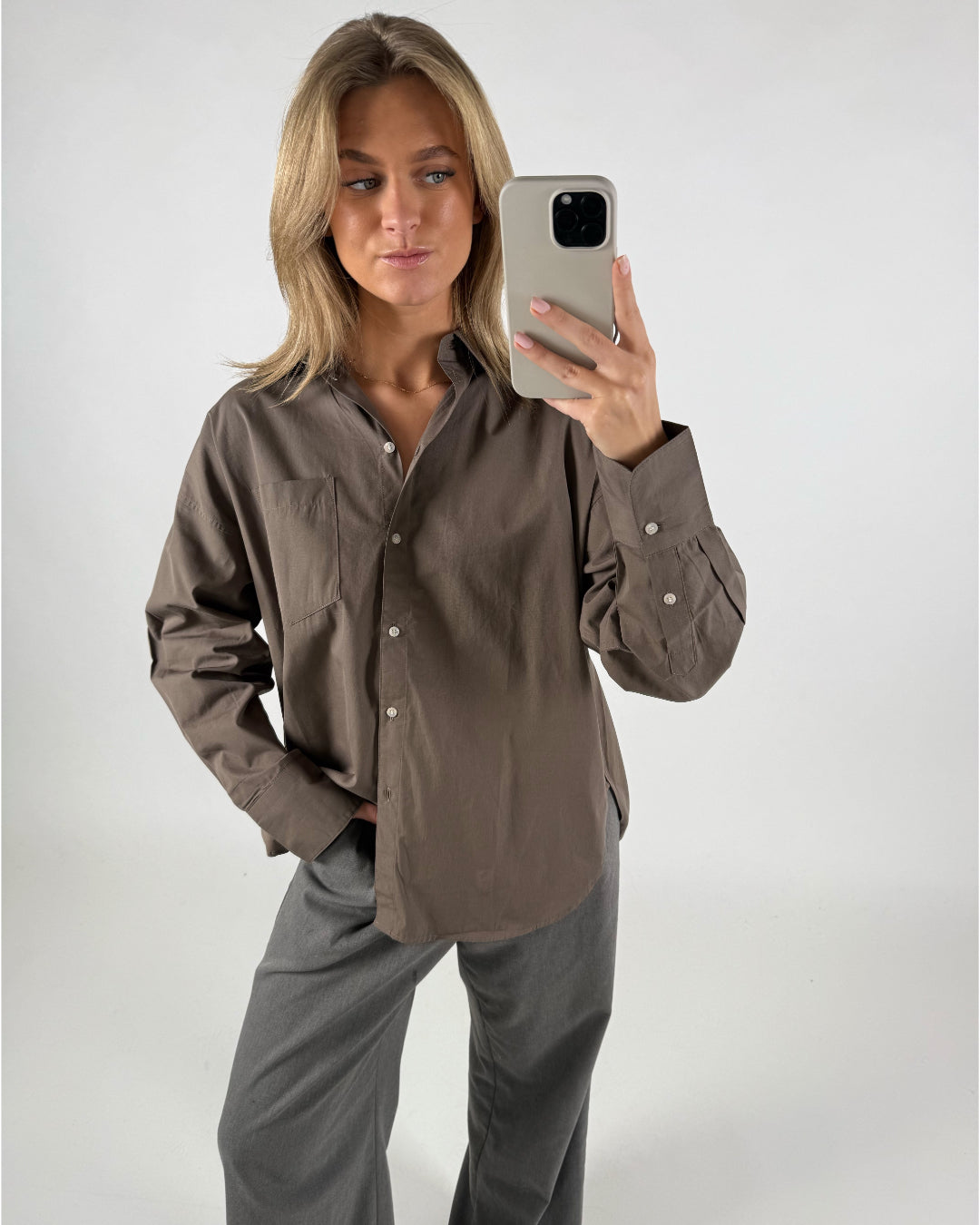 Kvinde tager selfie iført brun skjorte og grå bukser. Hun står i et neutralt, hvidt rum og ser til siden, mens hun holder en telefon med et lysegråt etui.