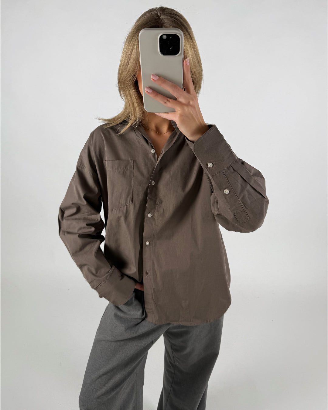 En person står iført en brun skjorte og grå bukser, holder en telefon op til ansigtet, i et minimalistisk, lysegråt rum.