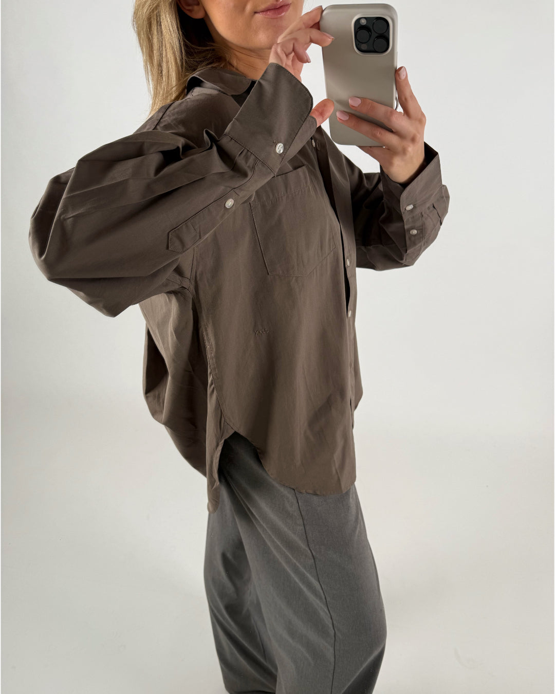 En person iført en stor brun skjorte og grå bukser tager et billede af sig selv med en smartphone i et neutralt, lyst rum.