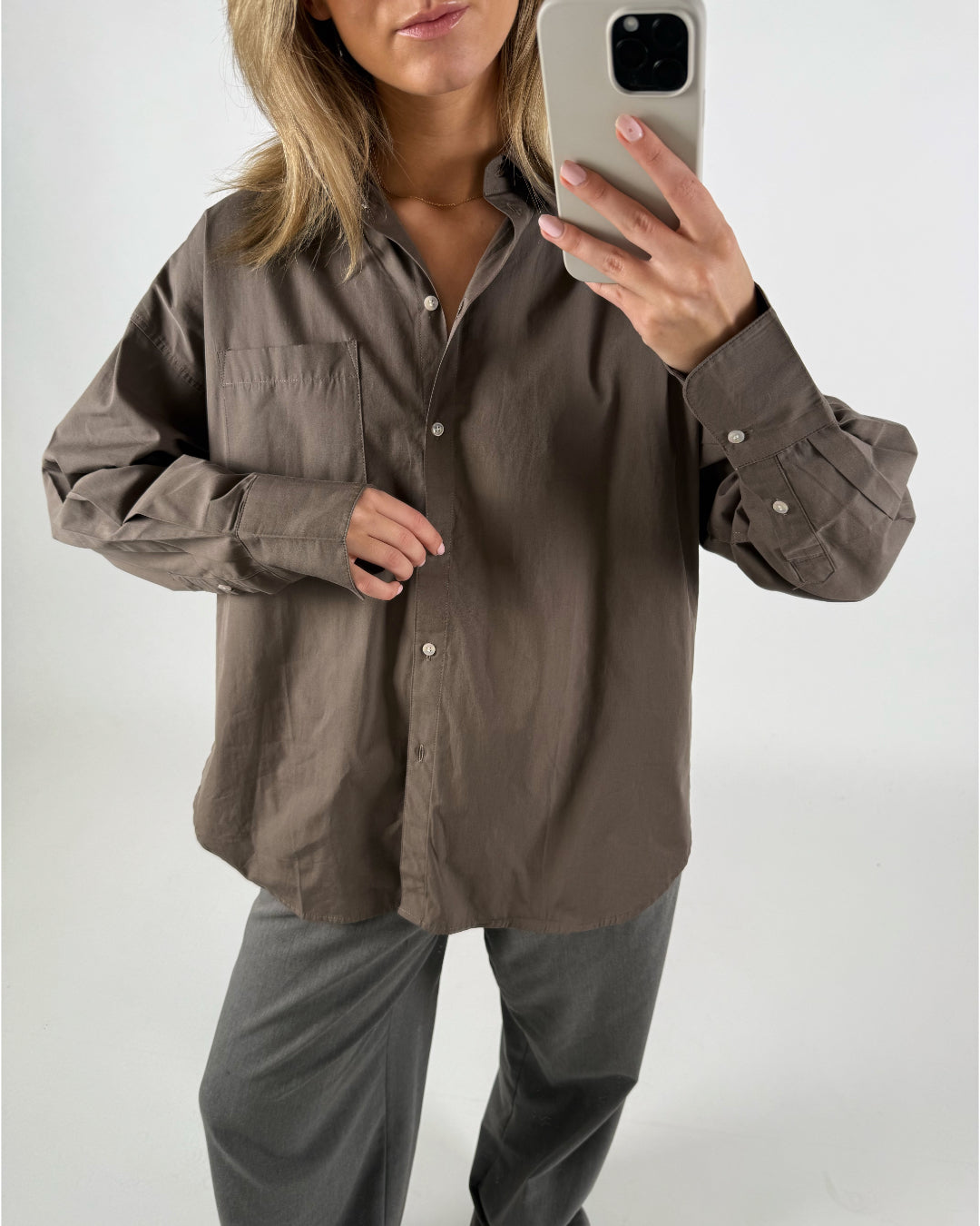 En person iført en brun skjorte tager et selfie med en smartphone. Personen står i et lyst rum med en neutral baggrund.