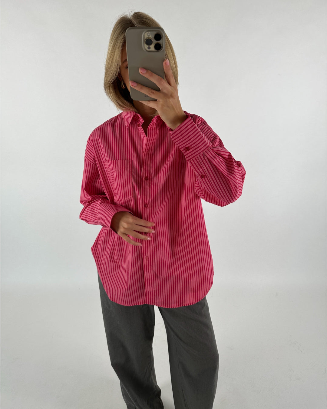 En person iført en pink stribet skjorte og grå bukser tager et billede af sig selv med en smartphone foran ansigtet i et hvidt rum.
