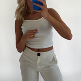 En person tager et spejl-selfie iført en hvid top og hvide bukser. Personen holder en blå telefon, og omgivelserne er minimalistiske med en hvid baggrund.