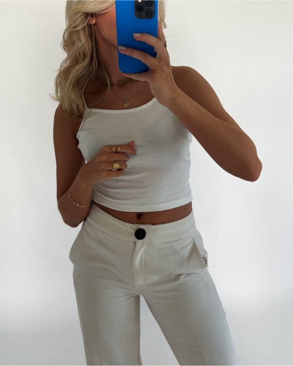 En person tager et spejl-selfie iført en hvid top og hvide bukser. Personen holder en blå telefon, og omgivelserne er minimalistiske med en hvid baggrund.