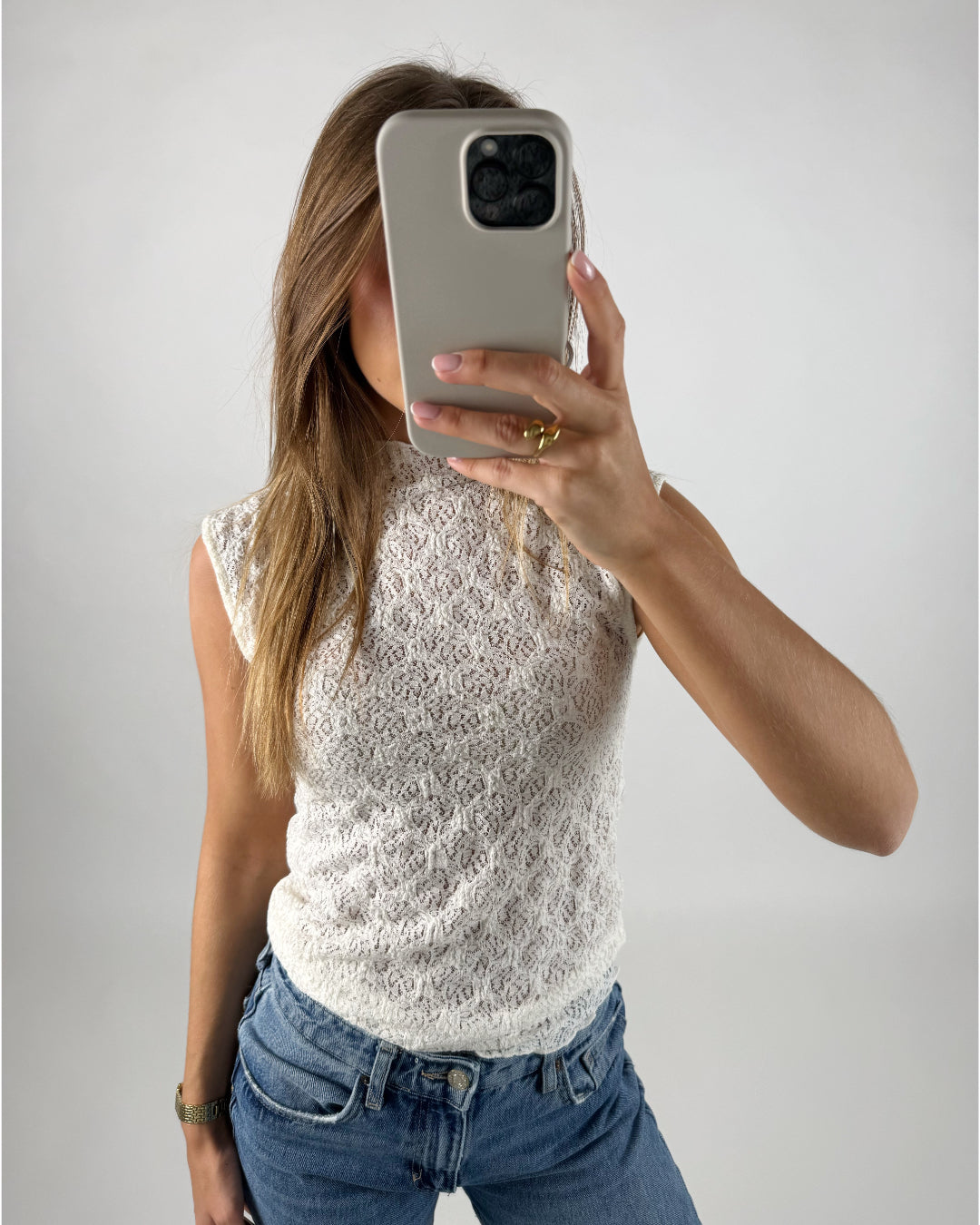 Personen holder en mobiltelefon foran ansigtet, iført en hvid blondetop og blå jeans, i et minimalistisk, hvidt rum.