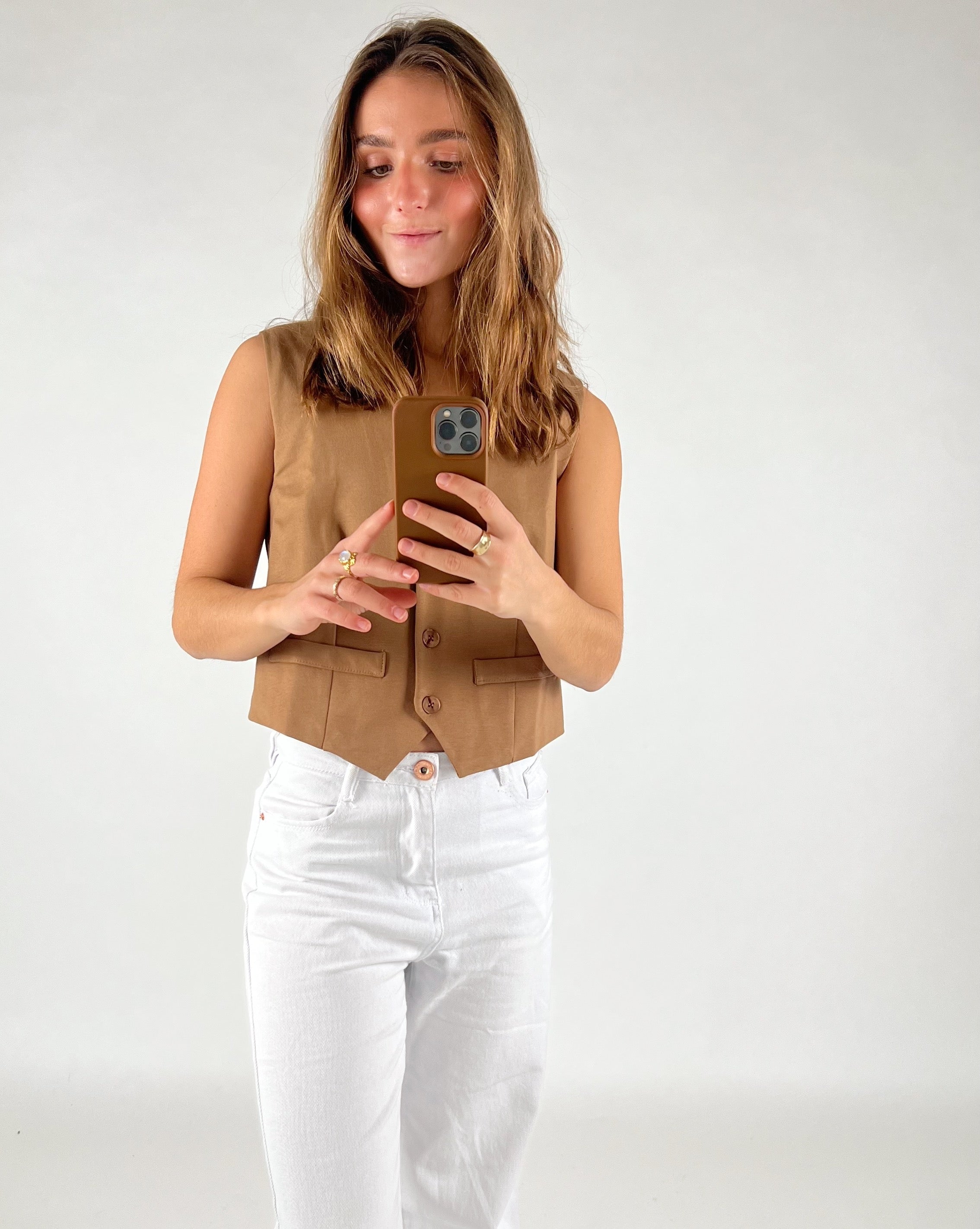 En person tager et billede med en smartphone iført en brun vest og hvide bukser i et neutralt rum.