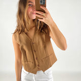 En person iført en brun vest tager en selfie med en smartphone i et lyst rum, iført hvide bukser.