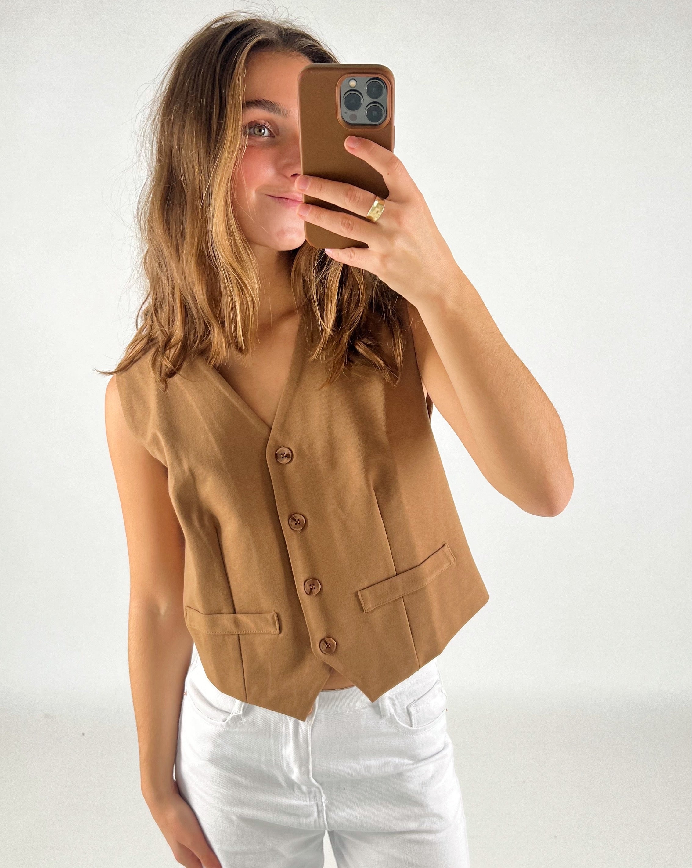 En person iført en brun vest tager en selfie med en smartphone i et lyst rum, iført hvide bukser.