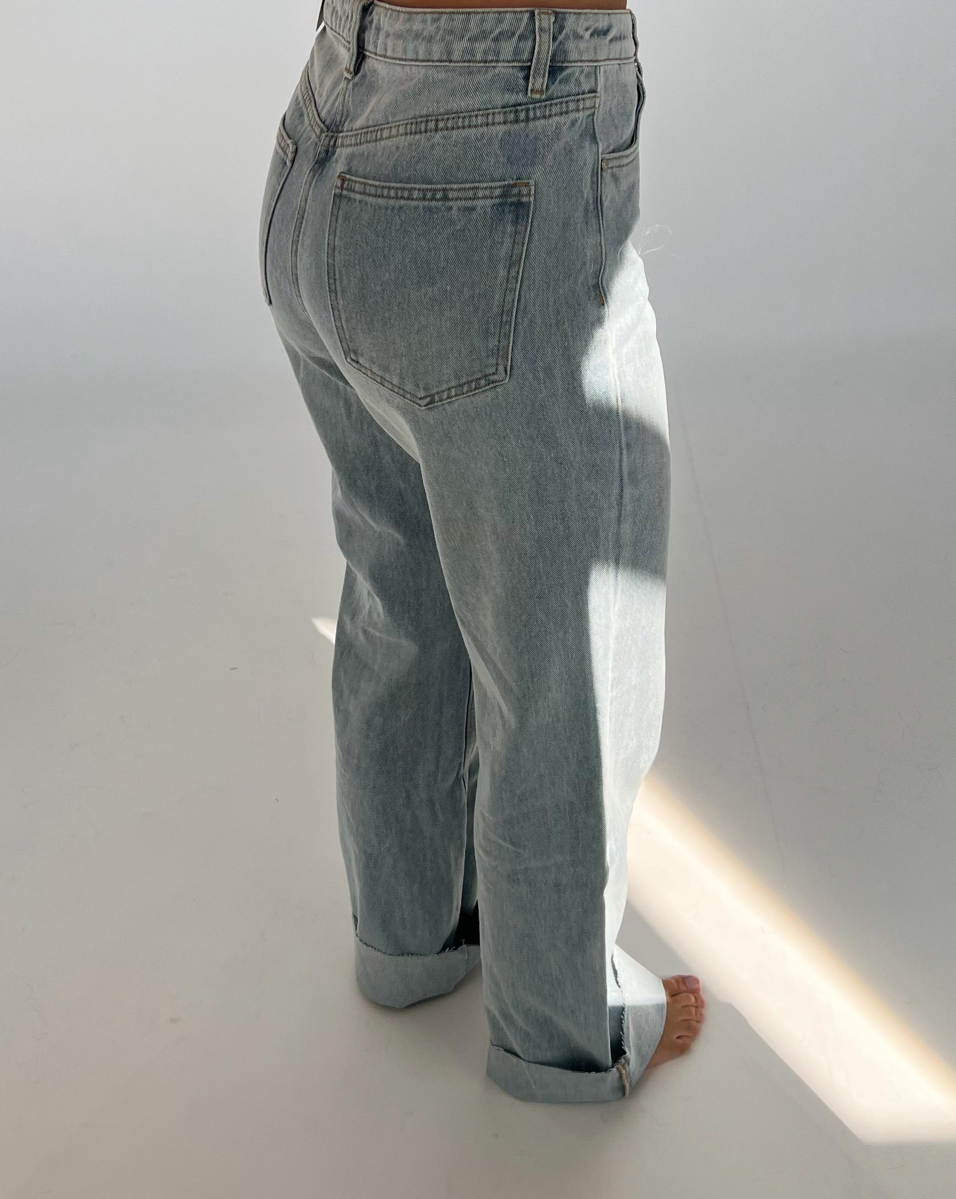 Bukserne er lyseblå denim, set bagfra, mens de bæres i et minimalistisk rum med hvid baggrund og skygger. Bunden af bukserne er let opsmøget.