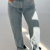 Person iført lyseblå jeans står med hænderne i lommerne i et lyst rum. Bunden af bukserne er rullet op, og der er solskin på den ene side.