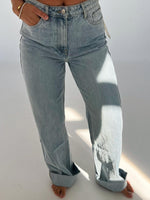 Person iført lyseblå jeans står med hænderne i lommerne i et lyst rum. Bunden af bukserne er rullet op, og der er solskin på den ene side.