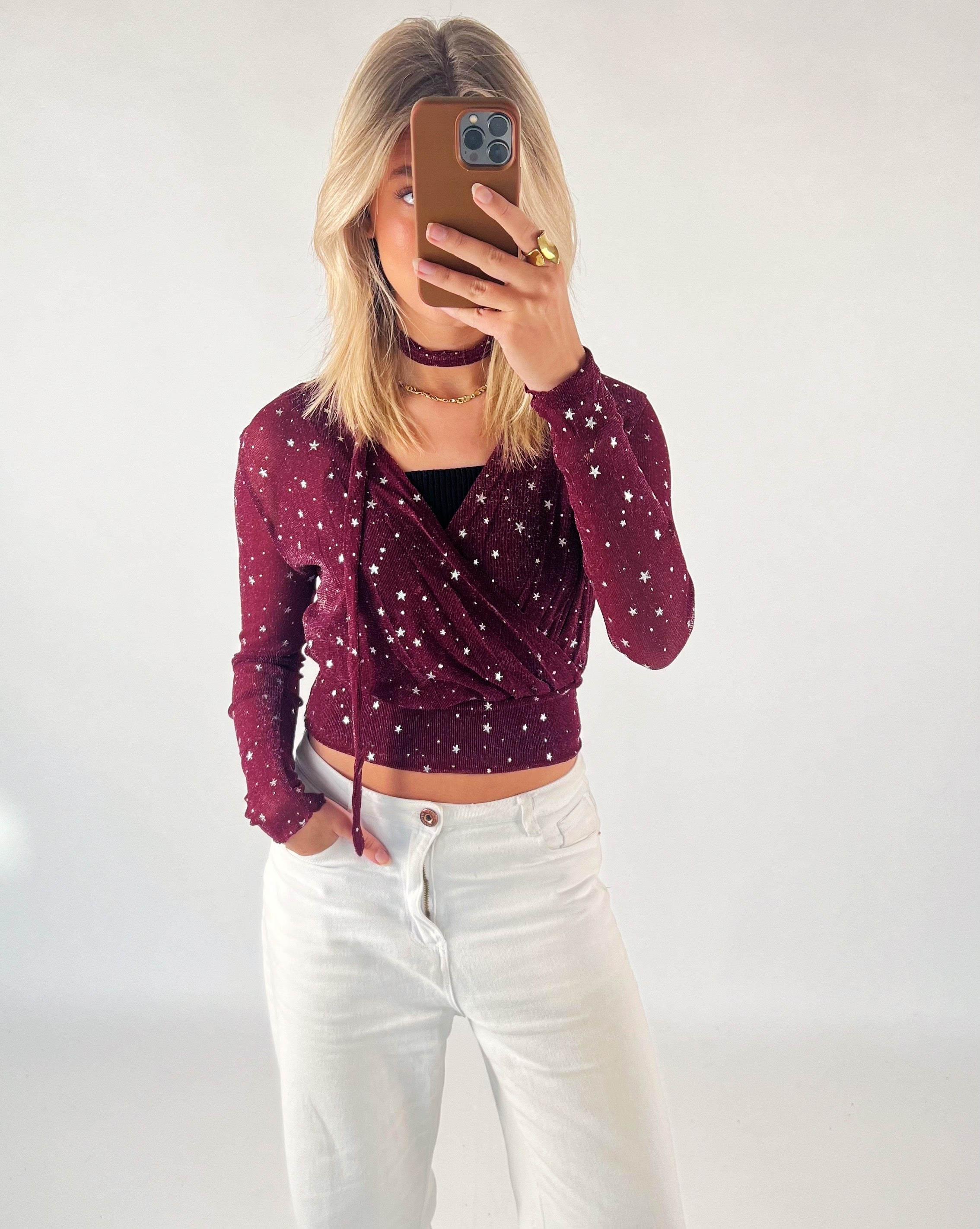 Person tager selfie, iført rød bluse med stjerner og hvide bukser, i et lyst rum.