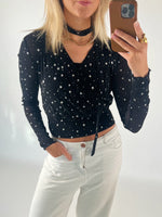 En person iført en sort stjernemønstret bluse tager en selfie med en brun indkapslet mobiltelefon. Personen bærer et guldhalskæde og hvidt buksetøj. Baggrunden er neutral.